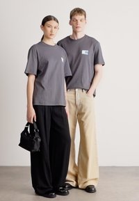 T-shirts cinzentas de algodão com um pequeno remendo colorido, calças largas pretas, calças de ganga claras e uma mala preta na mão do modelo.