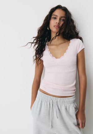 Jeune femme aux longs cheveux bouclés portant un haut côtelé rose clair avec bordure en dentelle et un pantalon de survêtement gris avec cordon, se tenant contre un mur blanc.