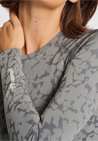 Camisa deportiva de manga larga en gris con un sutil patrón abstracto, hecha de una tela texturizada. Presenta un escote redondo y un detalle de logo en la manga.