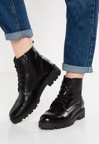 Stivaletti neri in pelle con lacci frontali, punta arrotondata e suola rigata. Cerniera laterale per facilitarne l'indossamento. Indossati con jeans blu arrotolati.