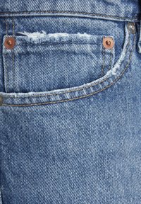 Jean en denim bleu présentant une poche arrière déchirée avec des bords effilochés, des rivets en cuivre et des coutures contrastantes le long des coutures.