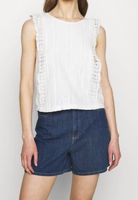 Vit ärmlös topp med spetsdetaljer längs axlarna, texturerade vertikala ränder och rak skärning, tillsammans med denimshorts.