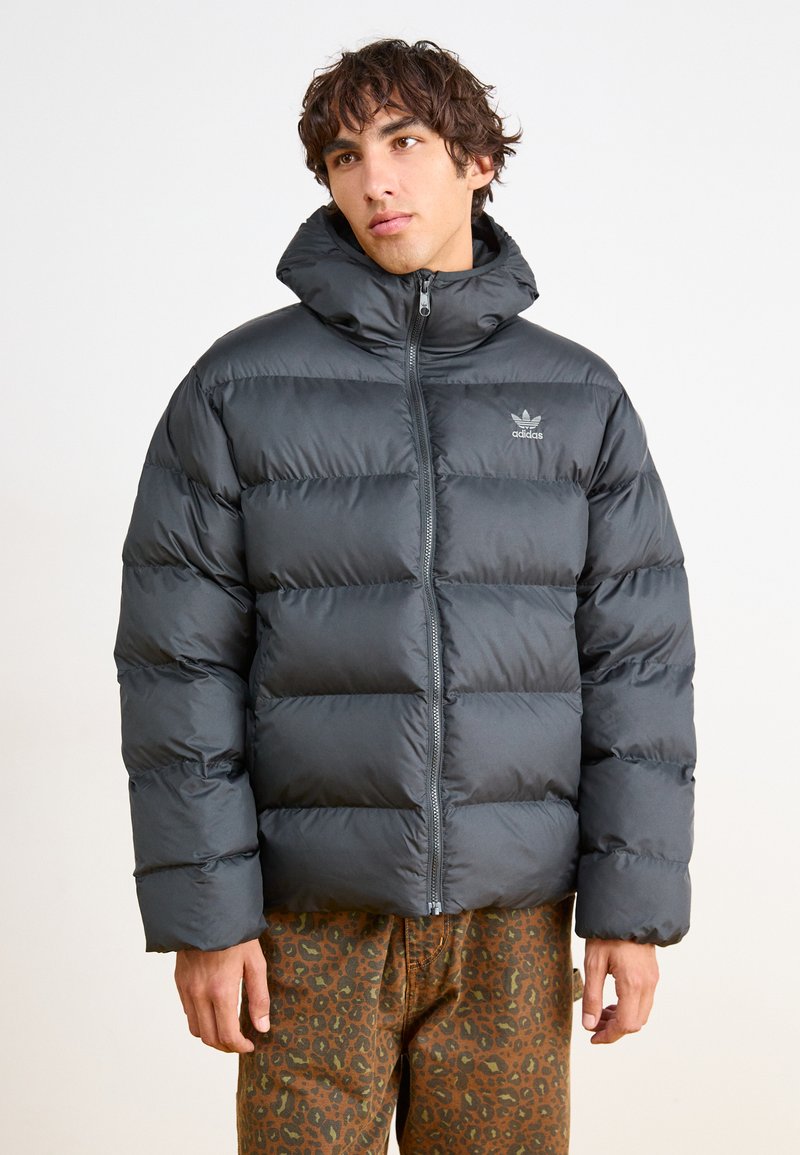 Schwarze Pufferjacke mit hochgeschlossenem Kragen, ausgestattet mit einem Reißverschluss vorne und einem ausgeprägten Steppmuster. Kombiniert mit braunen Leopardenzug-Print-Hosen.