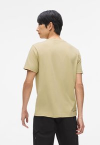 Krátky rukáv olivovo zeleného trička vyrobeného z mäkkej tkaniny, s okrúhlym výstrihom a jemným logom na leme, navrhnuté pre voľný strih.
