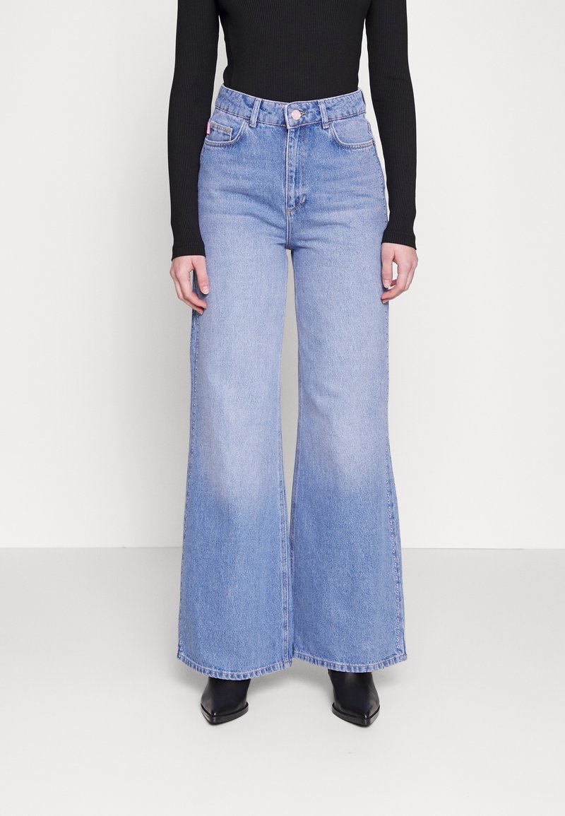 Persona che indossa jeans a vita alta, di un blu chiaro e a gamba larga, un top nero aderente a maniche lunghe e stivali neri con punta appuntita, in piedi davanti a uno sfondo bianco.