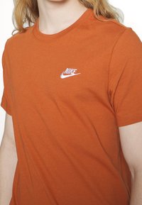 Oranžové bavlněné tričko s kulatým výstřihem a krátkým rukávem. Na levé hrudi je vyšitý bílý logo Nike.
