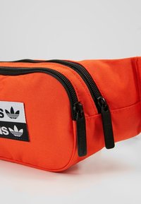 Orange fabric waist bag featuring two black zipper compartments, a textured surface, and a logo patch with three black stripes on a white background.