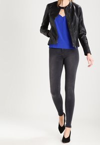 Schwarze Lederjacke mit hohem Kragen, über einem blauen Oberteil getragen, kombiniert mit eng anliegenden grauen Jeans und schwarzen High Heels. Glatte Textur, elegantes Design.