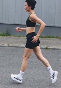 Soutien-gorge de sport noir et shorts, baskets Adidas blanches et chaussettes blanches. Mettez l'accent sur la posture athlétique de la coureuse et le design épuré et minimaliste.