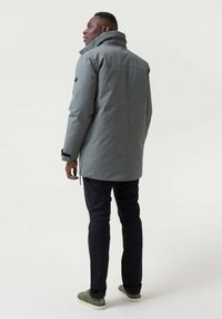 Chaqueta gris impermeable con alto cuello, puños elásticos y cierre delantero, combinada con pantalones negros y zapatillas verdes.
