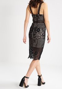 Musta pitsimekko, jossa on pilkkuprintti, muotoonommeltu siluetti, viistetyt hihnat ja polvenpituinen helma, jossa on kukkapitsikoristeita.