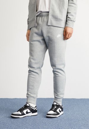Osoba ubrana w jasnoszare dresy Nike i dopasowaną bluzę z kapturem na zamek, stojąca na niebieskim dywanie w czarno-białych sneakersach Nike.