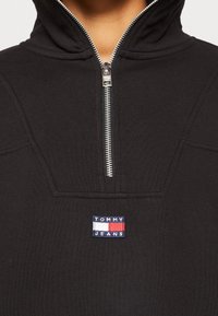 Felpa nera con zip argento, caratterizzata da una piccola patch logo "TOMMY JEANS" in blu, bianco e rosso sul lato del petto. Dettaglio in tessuto testurizzato.
