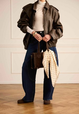 Person wearing oversized brown leather jacket, white turtleneck, wide-leg jeans, holding brown handbag with polka dot scarf in bright room.