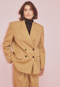 Woman with short brown hair wearing tan double-breasted blazer and pants, black turtleneck, and silver hoop earrings against pale pink background.