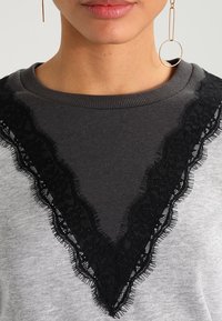 Sudadera gris con un borde de encaje negro que forma una forma de V en el escote. La tela tiene una textura suave y un detalle de diseño refinado.