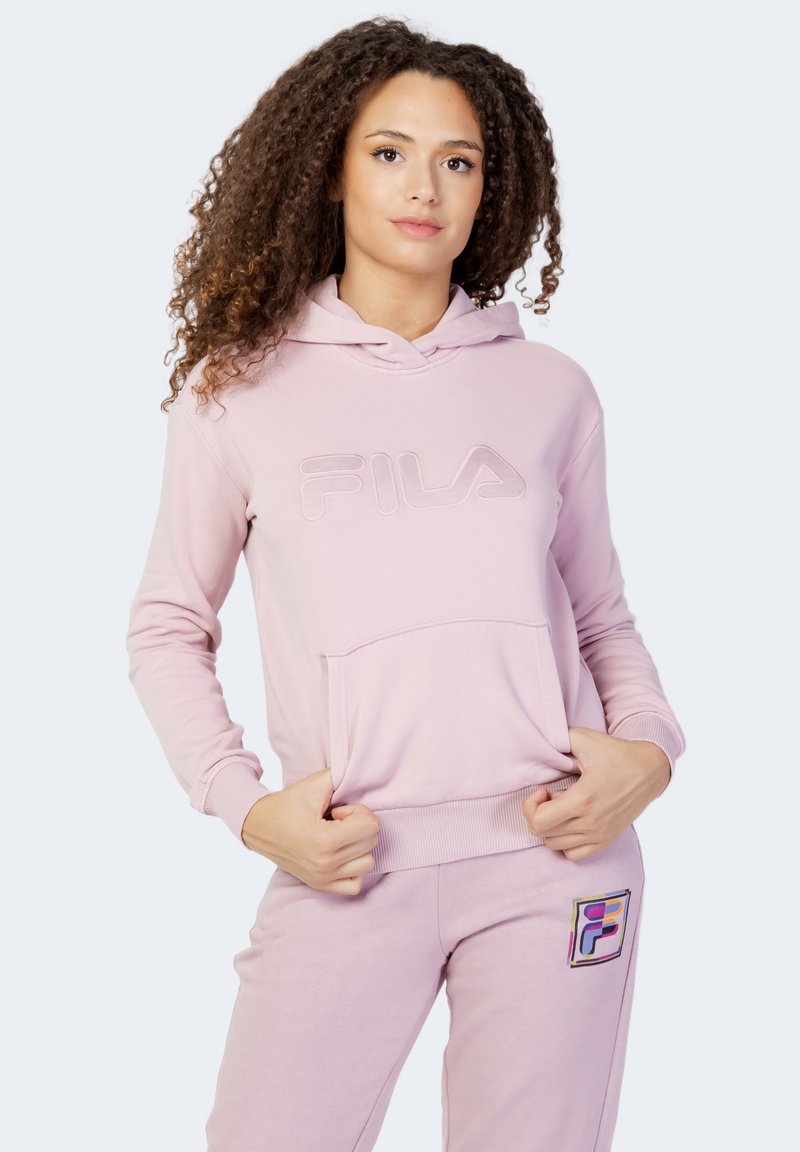 Felpa rosa chiaro realizzata in tessuto morbido, con tasca a sacca frontale e logo "FILA" ricamato. Abbinata a pantaloni tuta coordinati con una toppa multicolore.