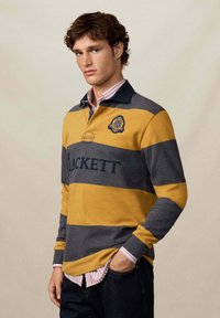 Mlađi muškarac s kovrčavom kosom nosi rugby majicu prugastu u senfu i sivoj boji s natpisom "HACKETT", preko ružičaste košulje s dugim rukavima koja također ima pruge.