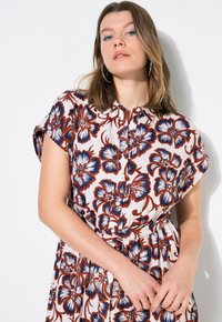 Vestido floral en crema con flores rojas y azul marino, mangas cortas dobladas, parte delantera con botones y una cintura ajustada acentuada por un cinturón.