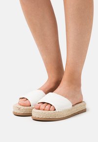 Sandales blanches à glissière avec un design matelassé, semelle en jute naturel, et fond en caoutchouc. Orteils découverts, texture lisse et talon ouvert.