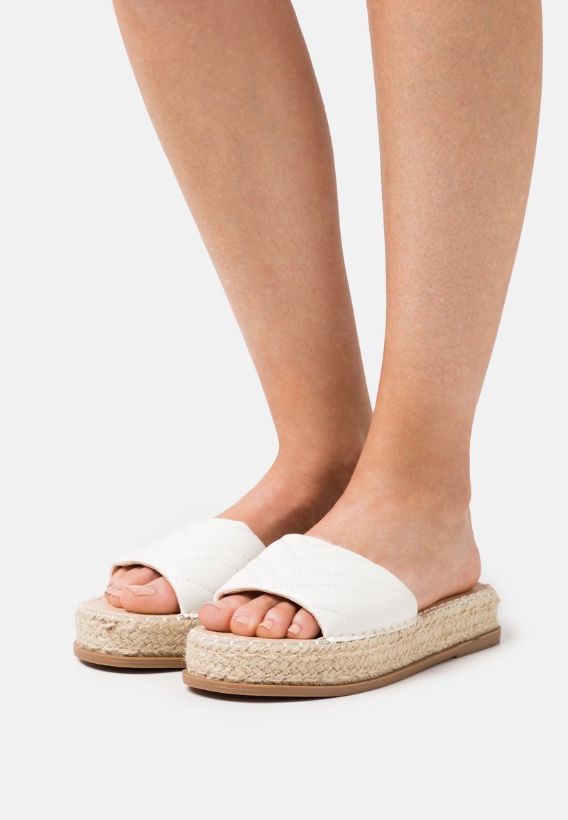 Sandales blanches à glissière avec un design matelassé, semelle en jute naturel, et fond en caoutchouc. Orteils découverts, texture lisse et talon ouvert.