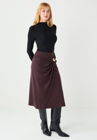 Black long-sleeve top paired with a knee-length burgundy skirt featuring a twisted design and gold hardware accent, with knee-high black boots.