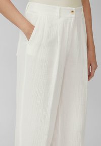 Pantalons blancs texturés avec une coupe décontractée, des poches latérales, une taille haute et une fermeture à bouton. Le tissu a une apparence froissée.