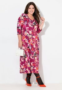 Vestido maxi floral en tonos de púrpura, rosa y naranja; cuenta con mangas 3/4 y un escote en V. Combinado con unos stilettos naranjas y un bolso blanco.