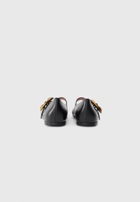 Ballerines en cuir noir avec une pointe arrondie, ornées d'une boucle dorée décorative et d'un petit talon. Texture lisse et design minimaliste.