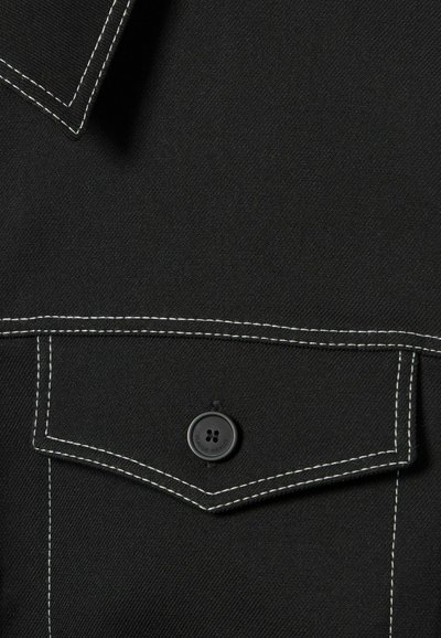 Tessuto nero con taschino sul petto e bottone nero, evidenziato da cuciture bianche a vista. Texture liscia e design classico da camicia.