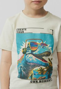 Crème-kleurig T-shirt met een kleurrijke dinosaurusafbeelding met zonnebril en koptelefoon, tekst luidt "CREËER JE EIGEN MOMENT!"