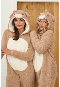 Twee vrouwen dragen bruine, fluffy onesies met witte accenten en capuchons in de vorm van een beer. De stof is zacht en gestructureerd, met ronde oren en gezichtsdetails.