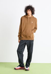 Ungdom som står med hånden i lommen iført brun Nike hettegenser, svarte bukser og hvite-røde sneakers foran en ensfarget bakgrunn.