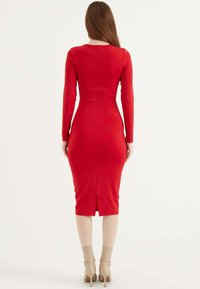 Czerwona dopasowana sukienka z długimi rękawami, sięgająca do połowy łydki, z rozcięciem z tyłu. Gładki materiał z elegancką sylwetką. Neutralne botki za kostkę.
