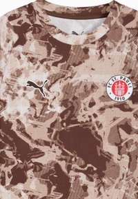 Brūna un beiga rakstaina sporta krekls ar īsām piedurknēm, apaļu kakla izgriezumu, ar Puma logo un FC St. Pauli emblēmu.