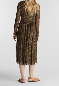 Långärmad, grön blommig klänning med en figursydd överdel och draperad midja, som flödar till knät; i kombination med bruna sandaler med remmar.