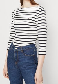 Vit långärmad topp med marinblå ränder, med en båtringning och en liten logga på bröstet, kombinerad med mörkblå högmidjade jeans.