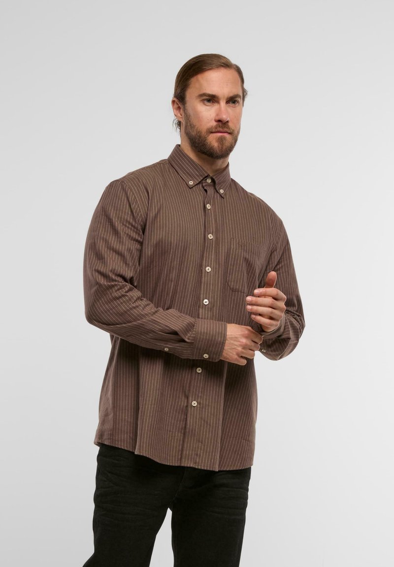 Chemise brune à manches longues, à boutons, avec des rayures verticales, un col pointu et une poche poitrine, accompagnée de pantalons sombres.