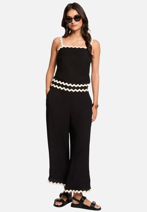 Kvinde iført sorte bukser med vide ben og matchende ærmeløs top med hvid bølget kant, sorte sandaler, solbriller og armbånd.