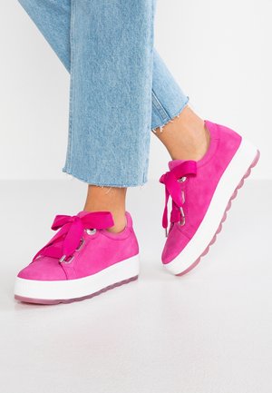 Pieds chaussés de baskets rose vif avec des semelles blanches épaisses et de larges lacets roses, associés à un jean bleu clair coupé court.