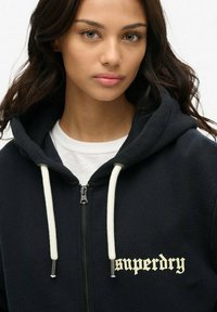 Sudadera con capucha de color azul oscuro, hecha de un material suave, con cremallera frontal, cordones blancos y texto de logo en color crema en el área del pecho izquierdo.