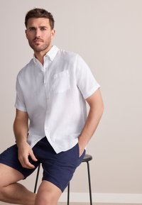 Chemise blanche à manches courtes en lin léger avec col boutonné et poche poitrine gauche, associée à un short bleu marine, assis sur un tabouret.