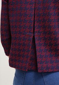 Navy and red patterned top, featuring textured fabric with geometric design and a back slit. Paired with blue denim.