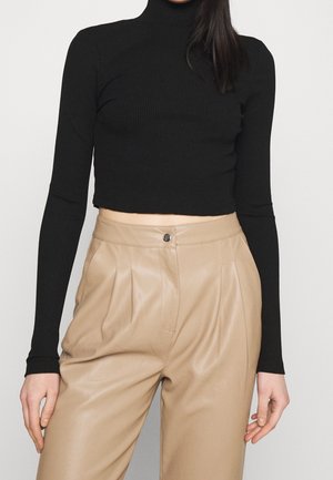 Mujer con un crop top negro de manga larga de canalé y pantalones de cuero beige plisados de talle alto sobre un fondo liso.