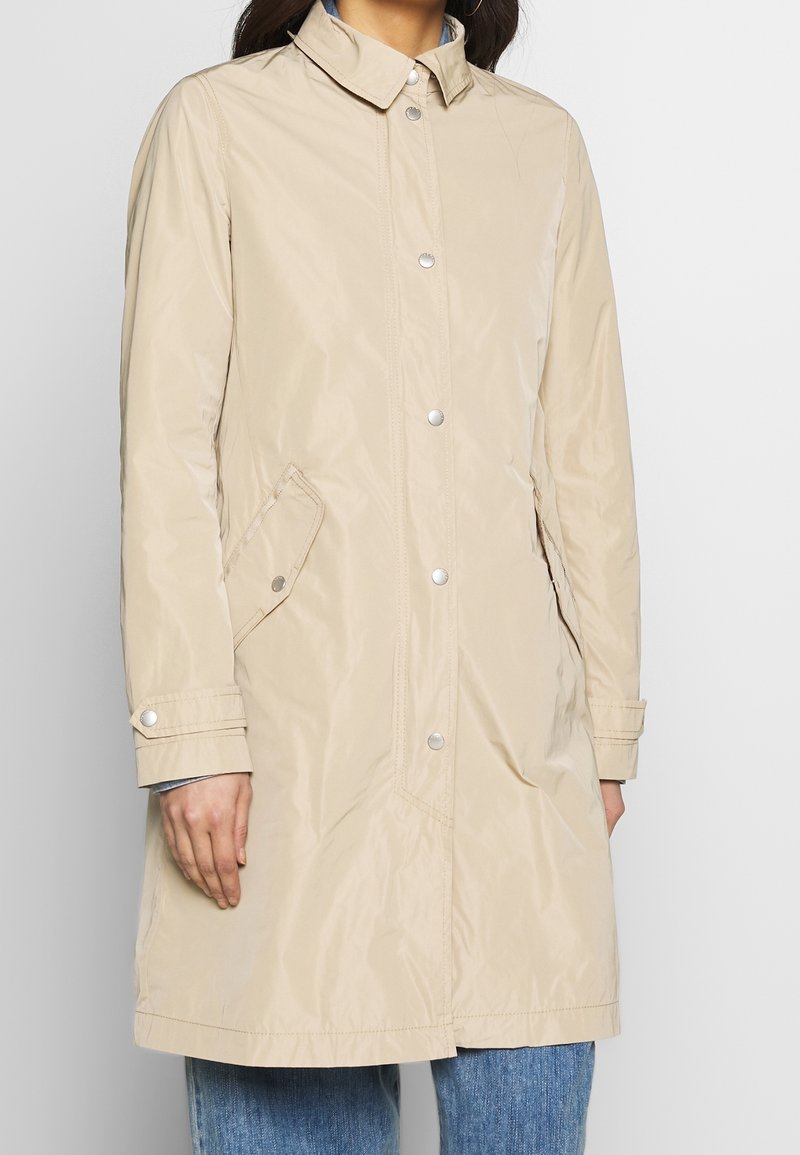 Manteau imperméable beige avec fermeture à boutons, col mao, poches latérales et coupe décontractée, présentant une texture lisse et un design simple.
