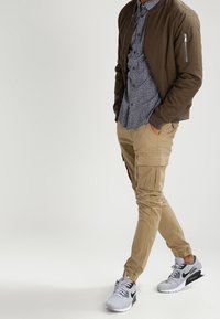 Bruine bomberjack, blauw met patroon overhemd, beige cargobroek met zakken, en grijze Nike sneakers met zwarte accenten. Details van een casual outfit zichtbaar.