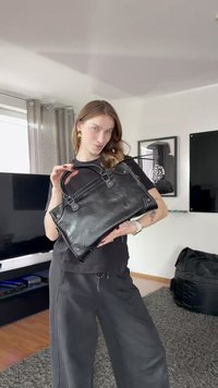 Fekete bőr kézitáska két fogóval, téglalap alakú, díszes fém kiegészítőkkel és texturált felülettel. Világos háttér előtt elhelyezve.