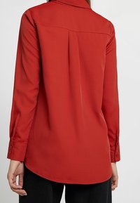 Chemise rouge à manches longues avec col, présentant une texture lisse et des poignets boutonnés. Le dos montre une empiècement et un ourlet incurvé subtil.