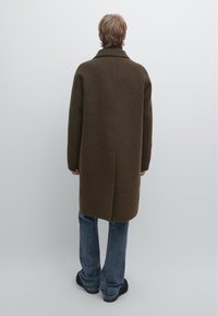 Manteau long en laine marron avec un col cranté, coupe droite et épaules tombantes ; porté sur un jean bleu évasé et des chaussures noires.