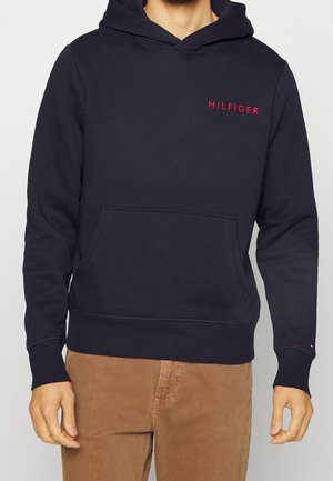 Homme portant un sweat à capuche bleu marine avec le texte "HILFIGER" en rouge sur la poitrine et un pantalon marron, debout devant un fond blanc.